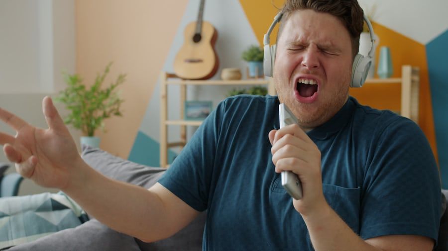 An adult man at home wearing headphones, eyes closed, singing into a phone held like a microphone