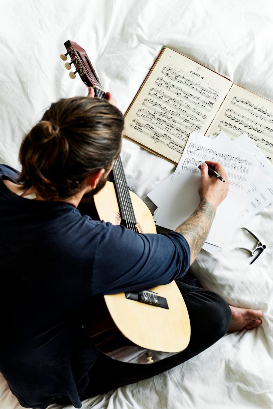 A guitarist sitting with an open score, loose sheets of manuscript paper and a pen, writing music out by hand