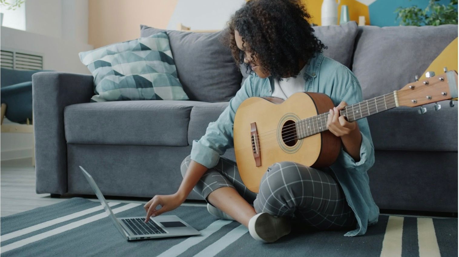 Person learning guitar online at home using a laptop while sitting on the floor