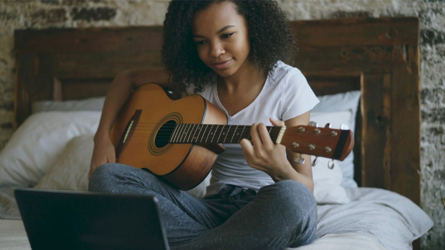 Woman practicing guitar on a bed while following an online lesson on a laptop