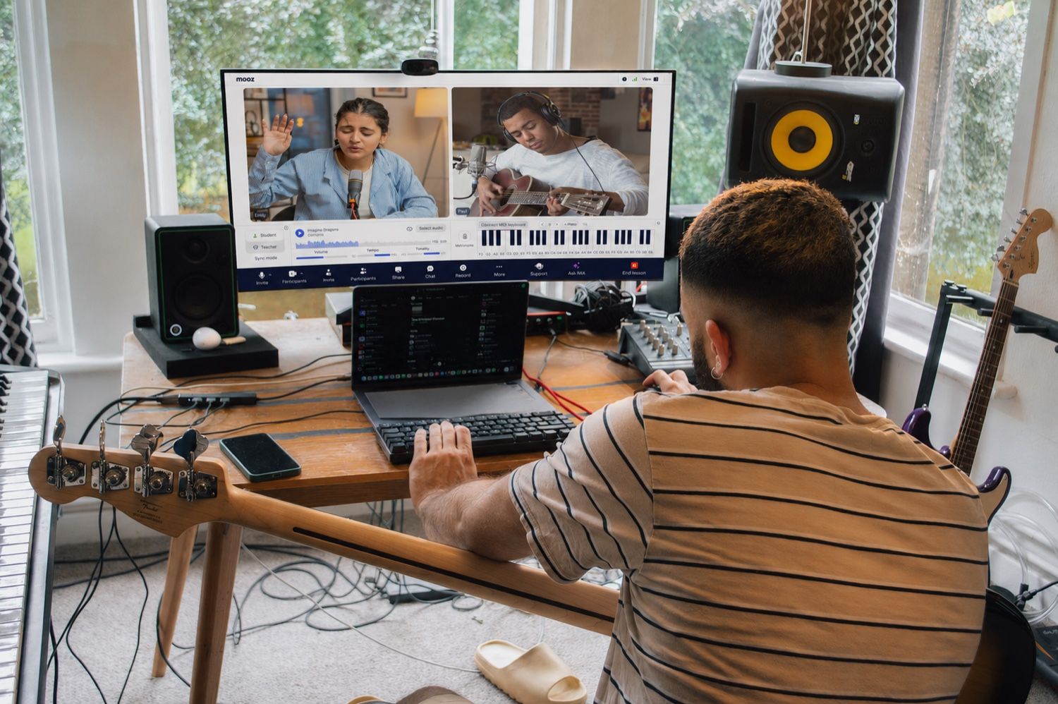 A music teacher at a home studio desk running an online lesson in MOOZ, with two video feeds on screen alongside Sync Mode, the audio player with tempo and tonality, a MIDI keyboard and a metronome