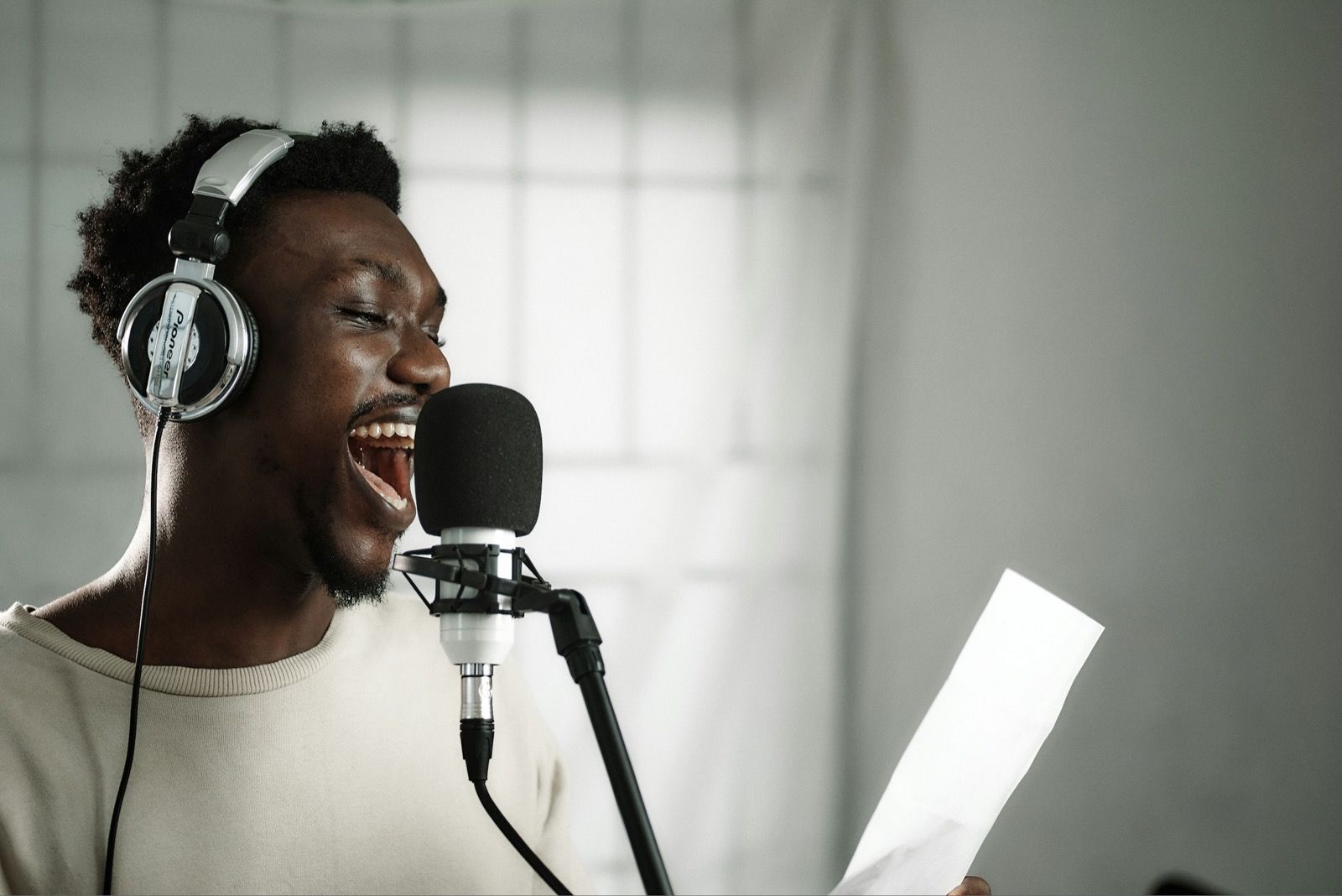 A singer wearing closed-back headphones, singing into a studio microphone while holding a lyric sheet